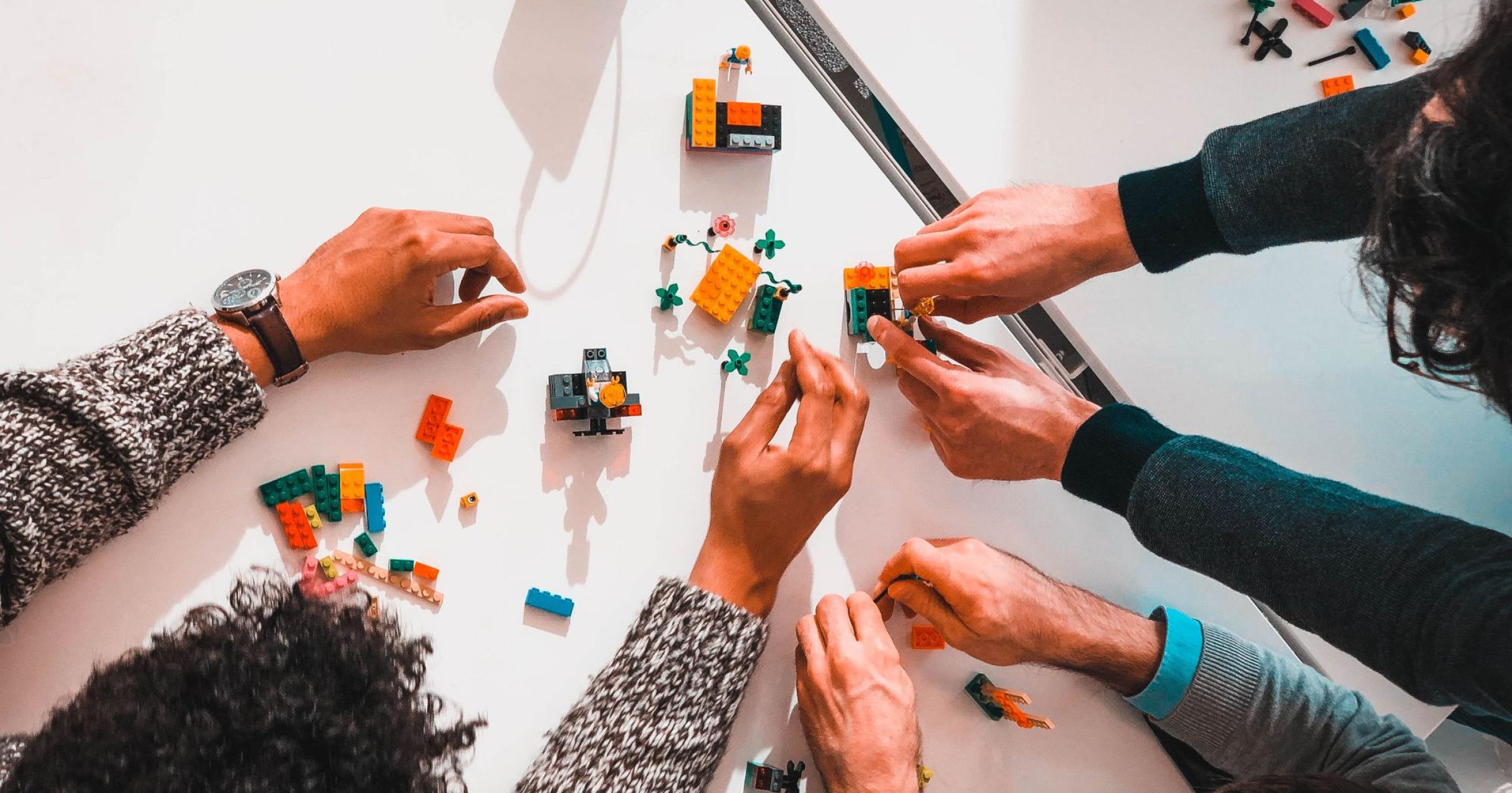 Top down photo of three people's hands working together to build things out of legos on a white table.
