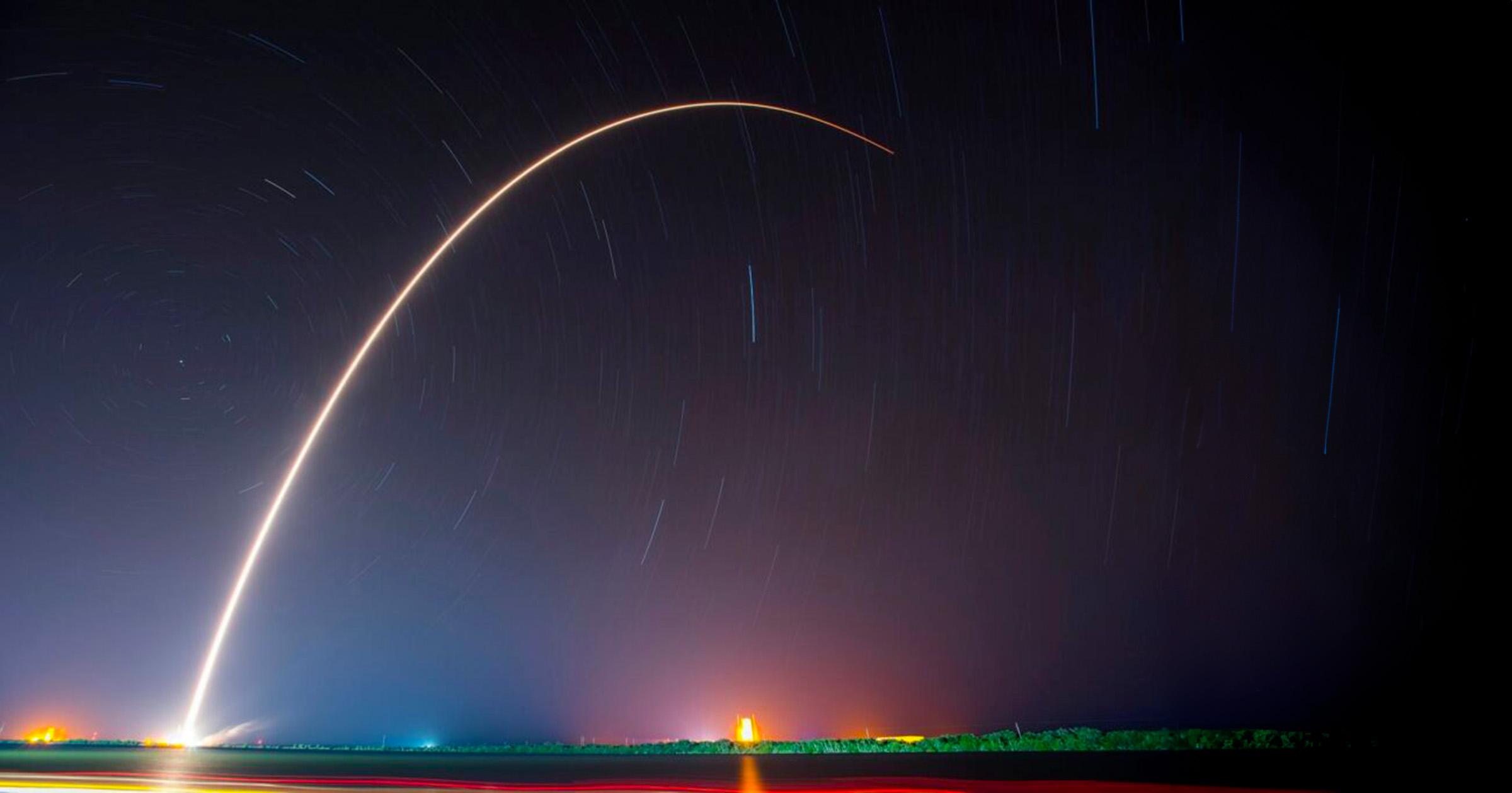 A night sky with stars streaking in the background and a streak of a rocket trailing in a large arc.